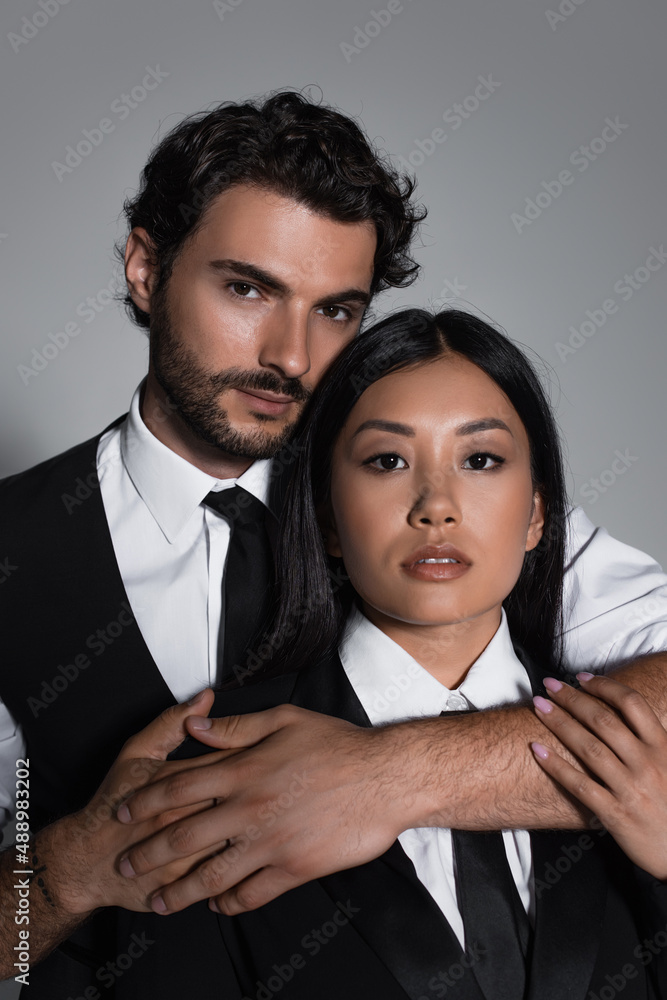brunette man embracing sensual asian woman while looking at camera isolated on grey brunette man embracing sensual asian woman while looking at camera isolated on grey