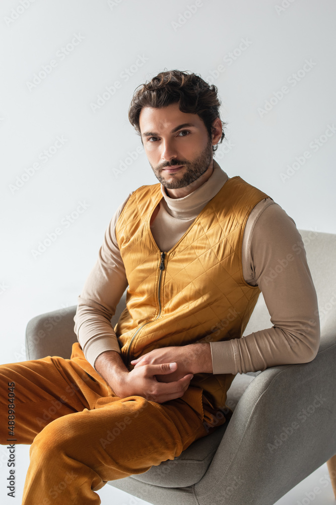 stylish man in yellow vest and trousers sitting in armchair and looking at camera isolated on grey stylish man in yellow vest and trousers sitting in armchair and looking at camera isolated on grey