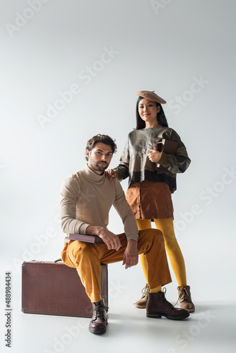 stylish asian woman holding books near man sitting on vintage suitcase on grey