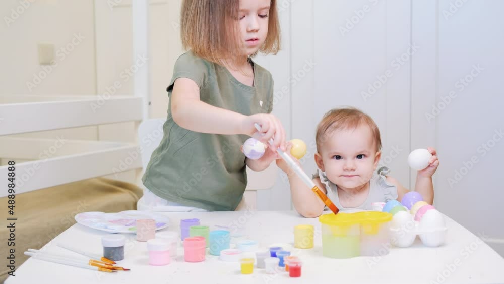 Preparation for Easter holiday. Older sister paints chicken eggs with paints. Baby with two milk teeth laughs, rejoices and has fun. Baby girl holds painted eggs in her hands and dances. Happy easter Preparation for Easter holiday. Older sister paints chicken eggs with paints. Baby with two milk teeth laughs, rejoices and has fun. Baby girl holds painted eggs in her hands and dances. Happy easter