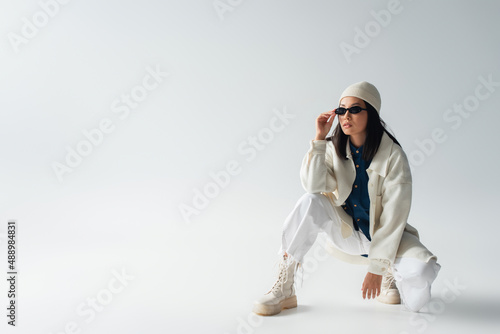 full length view of asian woman in white clothing adjusting sunglasses and looking away on grey