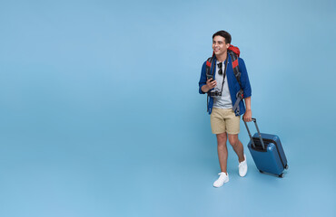 Happy young Asian tourist man holding smartphone with baggage going to travel on holidays isolated on blue copy space background.