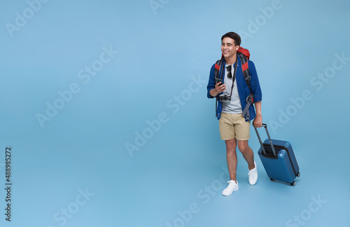 Happy young Asian tourist man holding smartphone with baggage going to travel on holidays isolated on blue copy space background.
