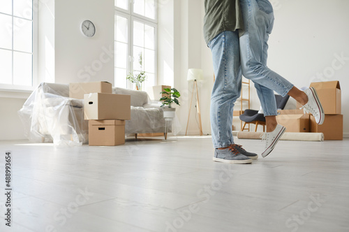 Happy married couple having romantic moment in new house. Young man and woman kissing while packing or unpacking boxes at home, cropped ground level shot of feet on floor. Buying real estate concept
