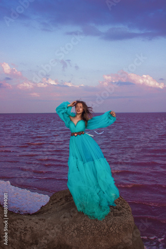 young girl with long hair in huge blue princess dress is standing with hand near head  and smiling cute near pink sea at sunset. travel concept, free space