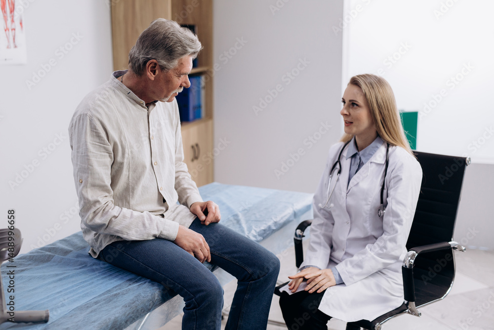 Obraz premium Grey haired male patient talking to female GP doctor. Senior man complaining about knee pain, explaining his health condition.