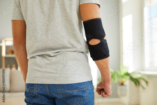 Young man wearing an adjustable black neoprene orthopedic elbow support brace on his right arm for easing pain in the elbow, back view, cropped close up shot. Concept of physical injury treatment
