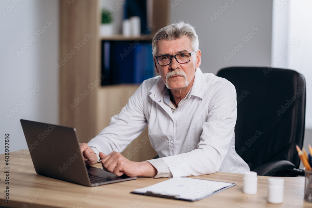 Serious male doctor sitting at workplace in hospital working on computer, looking thoughtful. Professional physician studying patient's electronic medical record. Serious male doctor sitting at workplace in hospital working on computer, looking thoughtful. Professional physician studying patient's electronic medical record.