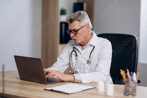 Serious male doctor sitting at workplace in hospital working on computer, looking thoughtful. Professional physician studying patient's electronic medical record.