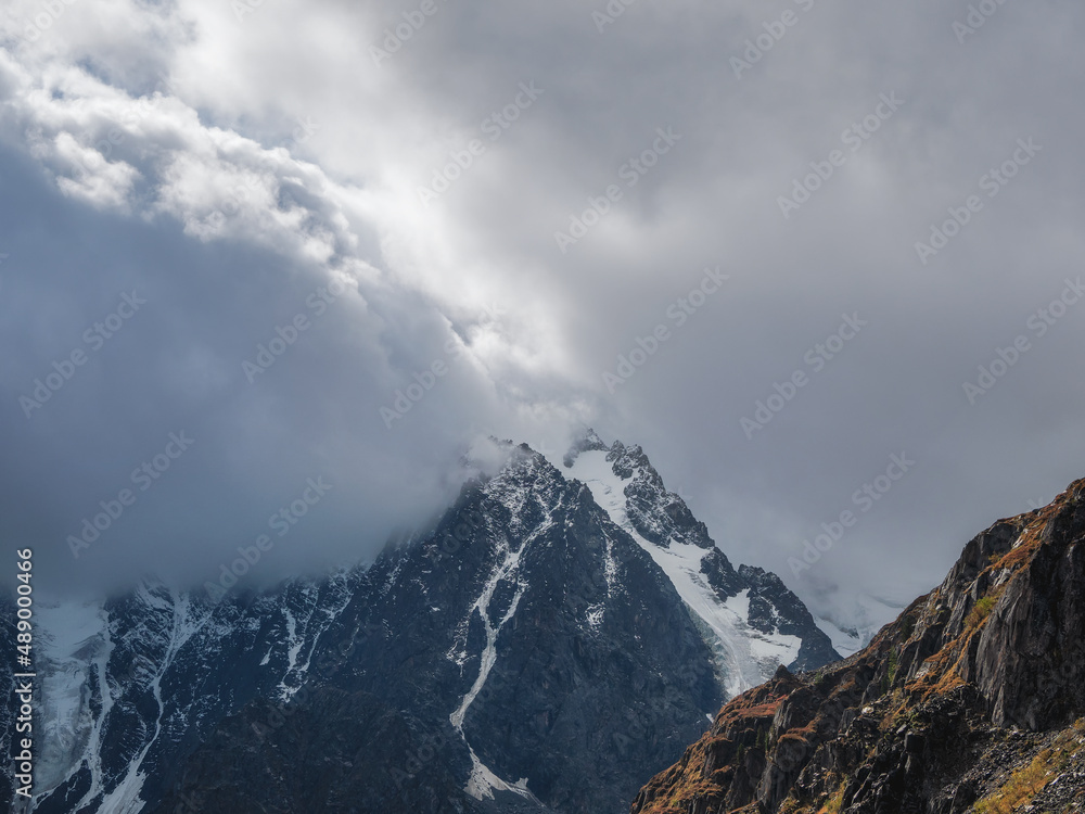 Dramatic aerial view to large snow mountain range under changeable rainy cloudy sky. Bright atmospheric alpine scenery with high snowy mountain peak in low clouds.