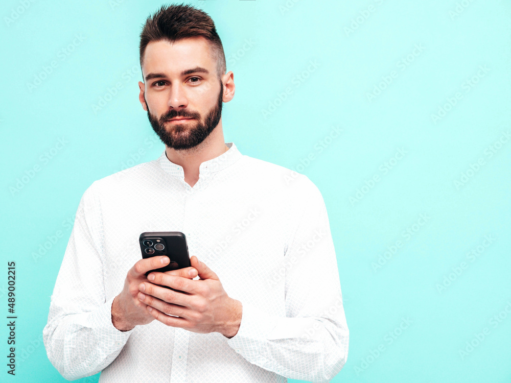 Handsome confident model.Sexy stylish man dressed in shirt and jeans. Fashion hipster male posing near blue wall in studio. Holding smartphone. Looking at cellphone screen. Using apps Handsome confident model.Sexy stylish man dressed in shirt and jeans. Fashion hipster male posing near blue wall in studio. Holding smartphone. Looking at cellphone screen. Using apps