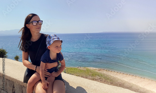A dark haired caucasian woman with a baby in her arms on vacation is sitting against the backdrop of beautifull azure sea water and mountains in a resort town