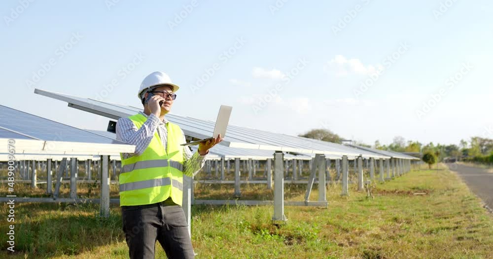 Slow motion shot, Young engineer in white helmet holding laptop computer in arm standing and talking on smartphone while working at solar farm in background Slow motion shot, Young engineer in white helmet holding laptop computer in arm standing and talking on smartphone while working at solar farm in background