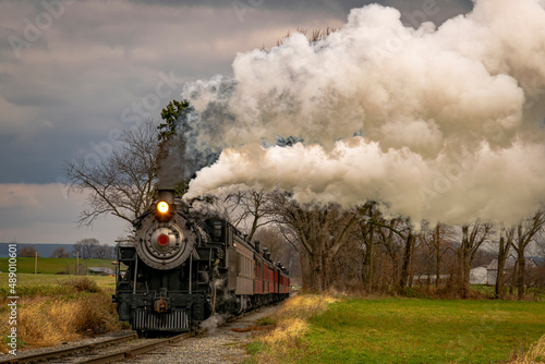 A View of an Antique Freight Steam Train Blowing Smoke Approaching Thru Trees in Late Afternoon on a Cloudy Day