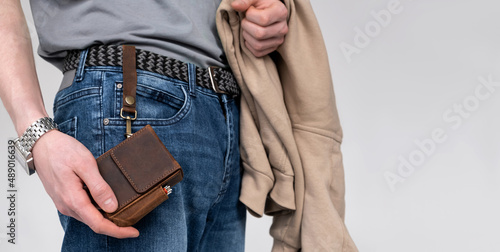 A man holds in his hand a cigarette case made of brown craft leather attached by a buckle to a belt on jeans on a light background. Horizontal orientation, no face, copy space. Stylish men's accessory photo