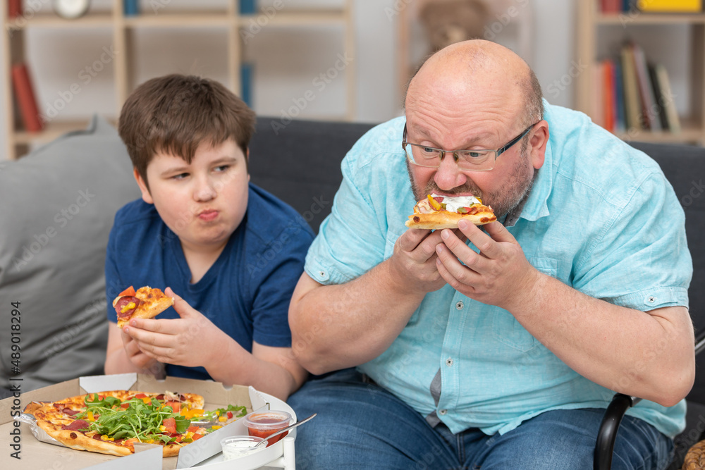 Dad and son are sitting on the sofa in the room eating pizza. Dad and son are sitting on the sofa in the room eating pizza.