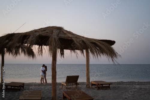 Young happy loving couple on the beach under straw umbrellas hugging at sunrise or sunset. Honeymoon.