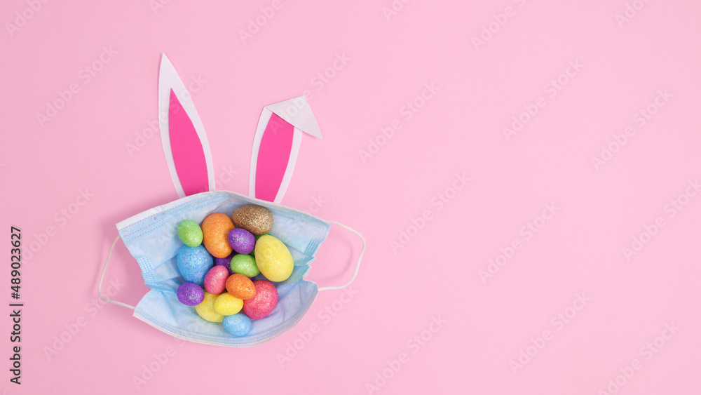 Face mask with colorful Easter eggs and bunny ears on pastel pink background. Creative copy space flat lay