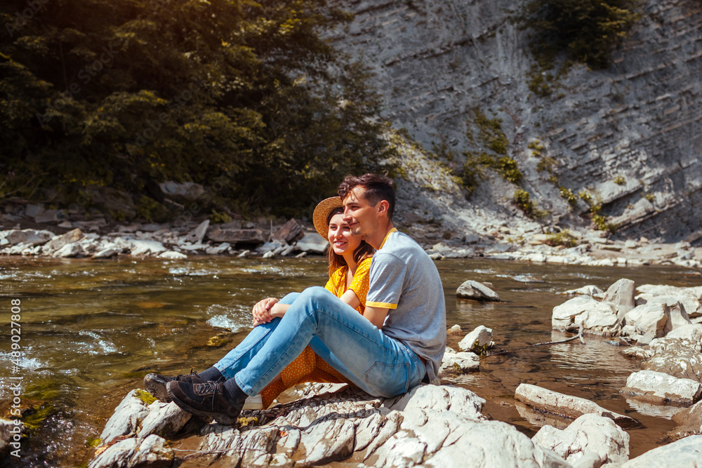 Couple of tourists relaxing by mountain river enjoying landscape. Travelers sitting on rock. Summer vacation