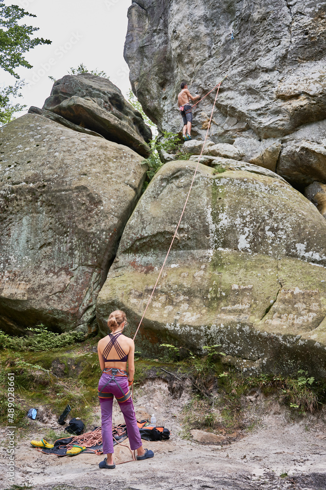 Female climber belaying leader during rock climbing outdoors, using rope. Back view of young woman looking to his rope partner. Concept of teamwork, trust, extreme sport and outdoor activity.