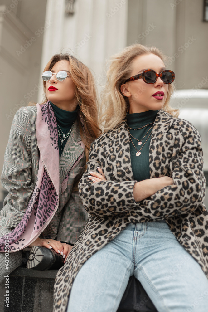 Fashion young pretty women with sunglasses in stylish clothes with leopard coat, blue jeans and gray suit sits on the street Fashion young pretty women with sunglasses in stylish clothes with leopard coat, blue jeans and gray suit sits on the street