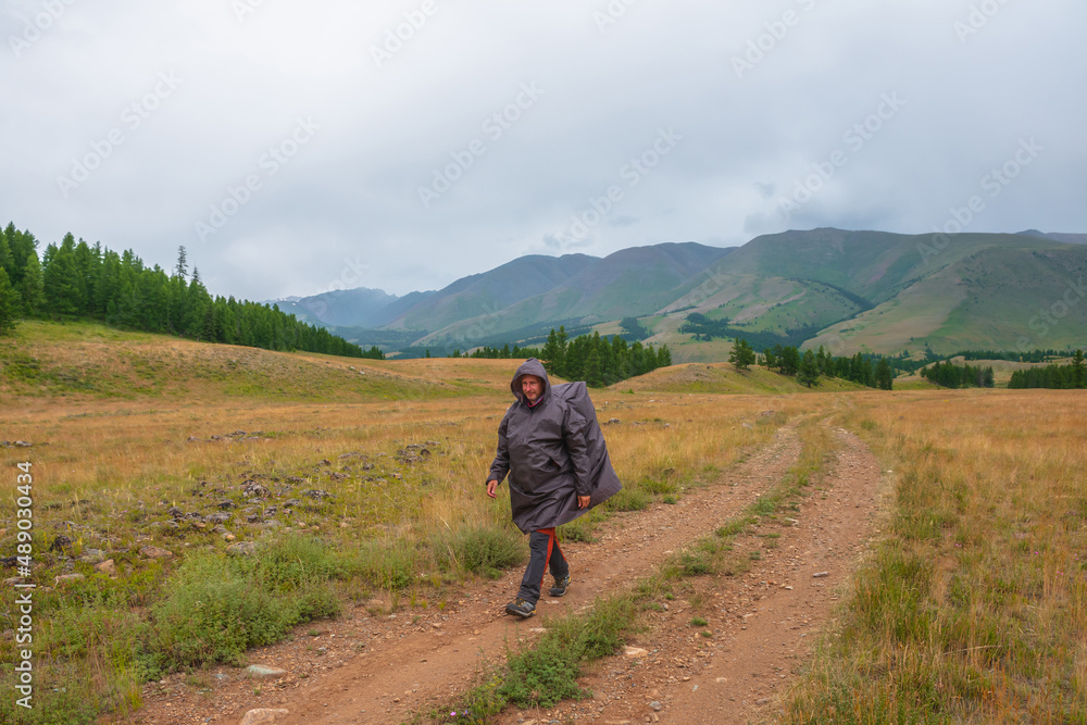 Satisfied tourist returns from mountains in overcast. Happy man in raincoat walks through hills and forest in bad weather. Traveler goes towards adventure. Hiker and mountain range under cloudy sky.