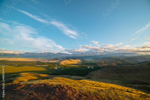 Scenic sunny view from sunlit grassy hill to forest valley with serpentine river against high snowy mountain range in sunlight. Beautiful snake mountain river in forest and snow mountains in sunshine.