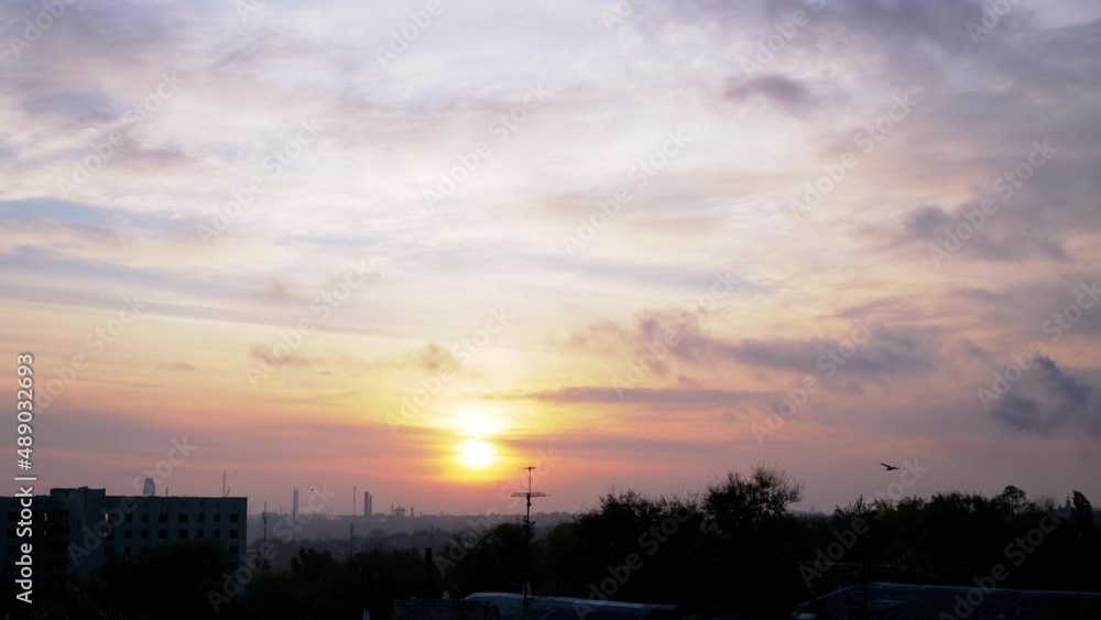 Sunrise Over a Sleeping Industrial City. Disappearing gray smoky clouds move over treetops and unfinished buildings. Rising soft sun over the red-orange horizon. Flying flock of birds. Early morning. Sunrise Over a Sleeping Industrial City. Disappearing gray smoky clouds move over treetops and unfinished buildings. Rising soft sun over the red-orange horizon. Flying flock of birds. Early morning.