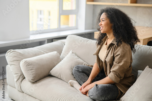Serene carefree young woman with long hair sitting at the sofa in modern apartment, looking away. Female resting at home, daydreaming, smiling