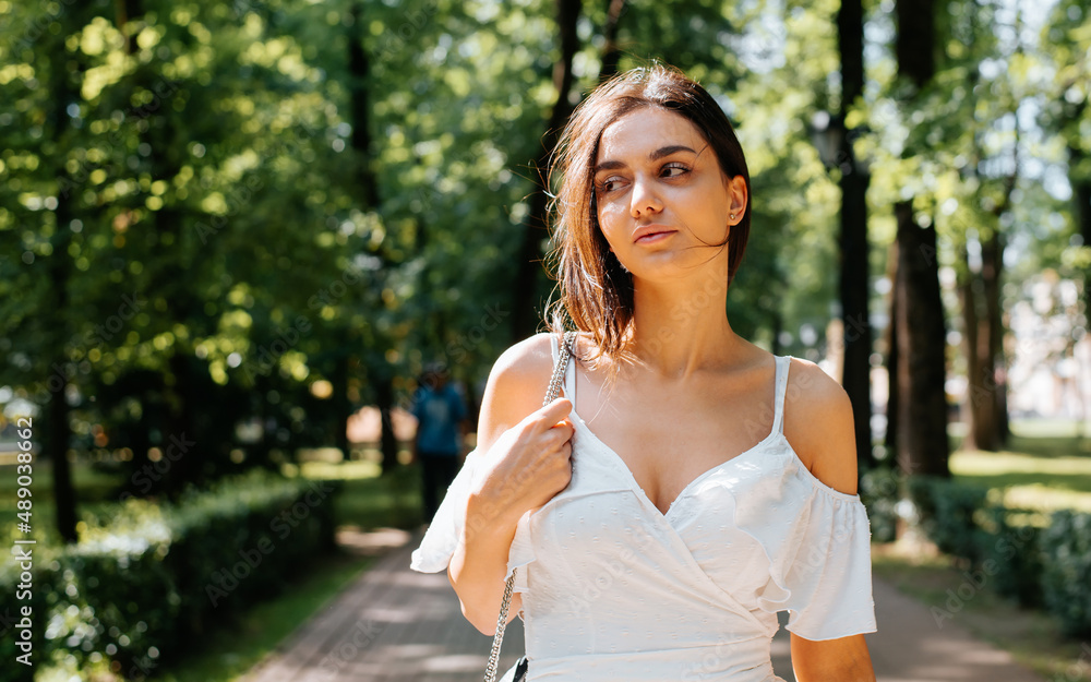 Pretty young indian woman in white summer dress walking on sidewalk in park on sunny day outdoors and looking away. Half-length portrait of beautiful mixed race brunette lady Pretty young indian woman in white summer dress walking on sidewalk in park on sunny day outdoors and looking away. Half-length portrait of beautiful mixed race brunette lady