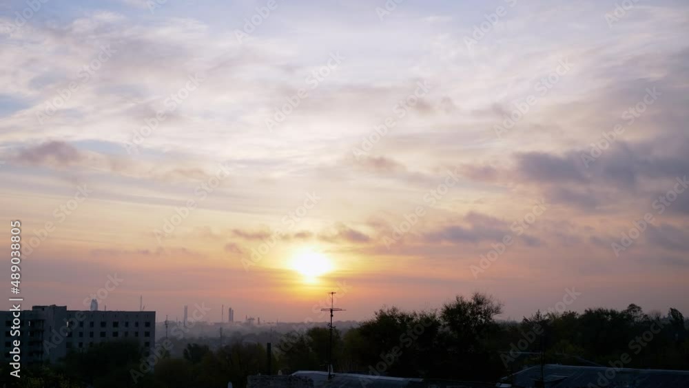 Sunrise Over a Sleeping Industrial City. Disappearing gray smoky clouds move over treetops and unfinished buildings. Rising soft sun over the red-orange horizon. Flying flock of birds. Early morning. Sunrise Over a Sleeping Industrial City. Disappearing gray smoky clouds move over treetops and unfinished buildings. Rising soft sun over the red-orange horizon. Flying flock of birds. Early morning.