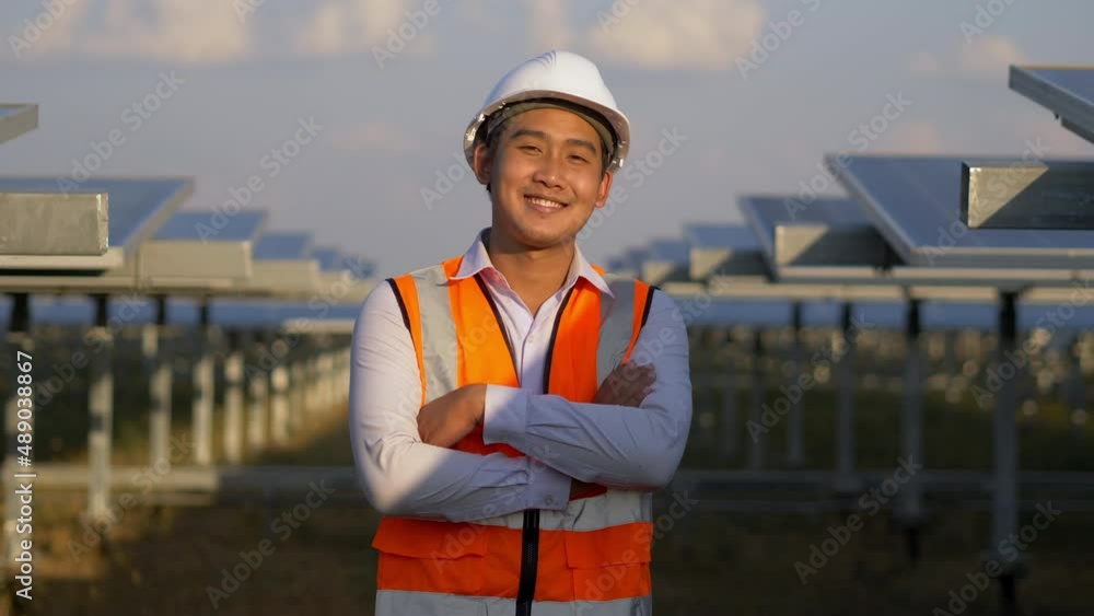 Dolly shot, portrait Asian young engineer wear white helmet standing and turn to looking to camera and crossing arms with smile at solar farm, solar panel in background Dolly shot, portrait Asian young engineer wear white helmet standing and turn to looking to camera and crossing arms with smile at solar farm, solar panel in background