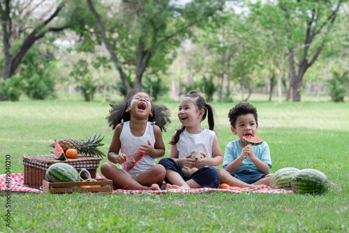 Happy little kids African, Caucasian and Asian picnic in the park. They laughing and sitting on grass eating watermelon with basket of fruits. Diverse ethnicity children friends enjoy life on summer © Pruksachat