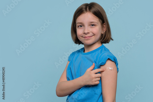 Child Vaccinated. Cute Little Girl pointing finger at adhesive plaster bandage on her arm after Being Injected Covid-19 Vaccine, light blue studio background. Children immunization. Health pass