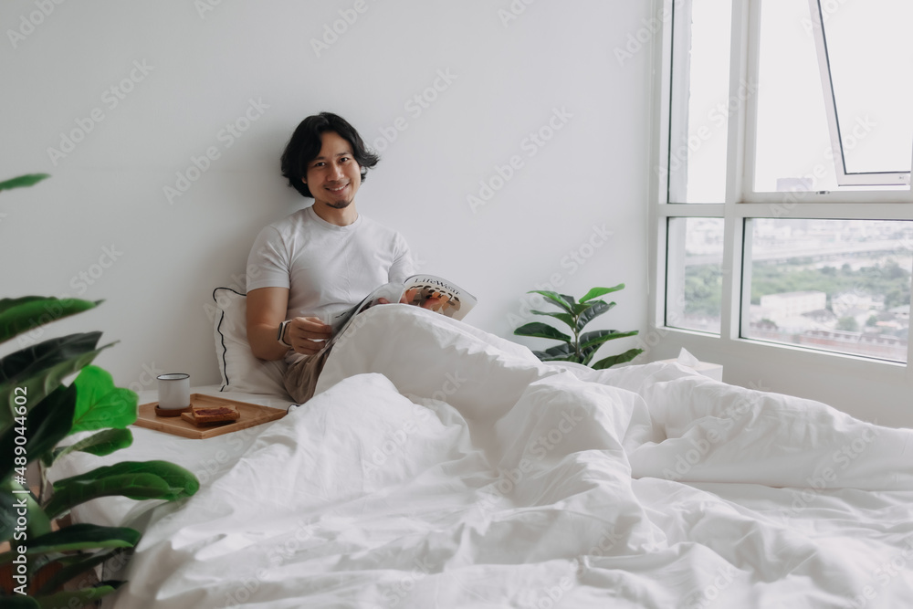 Asian man reading book on the bed with breads and coffee in his apartment.