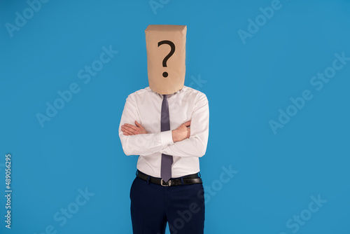 Businessman with brown paper bag on head with question mark, standing on blue studio background