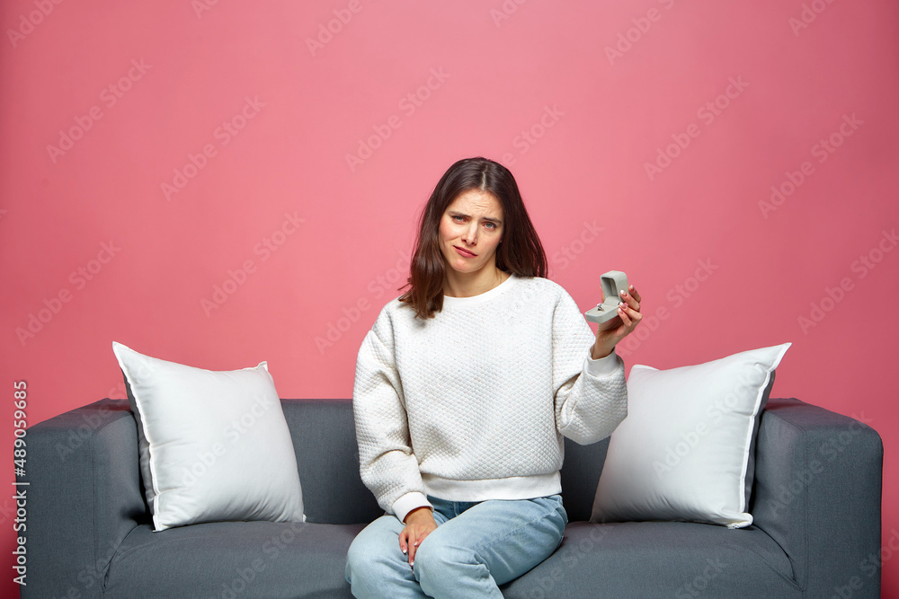 Upset young woman hold engagement ring received marriage proposal, grimacing, sitting on couch