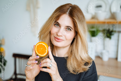 A pretty young white girl holds an orange slice in her hands. The blonde smiles at the camera. Citrus taste of orange fruit. Close-up portrait.