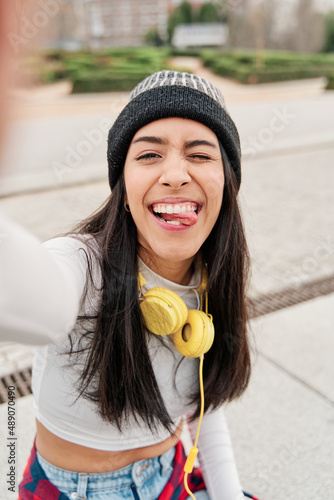 latina taking a selfie while making a funny face sticking out her tongue. Young Hispanic girl wearing casual clothes and a hat with headphones smiling. photo