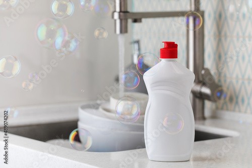 White bottle with dishwashing gel with soap bubbles around on the background of a sink with dirty dishes.