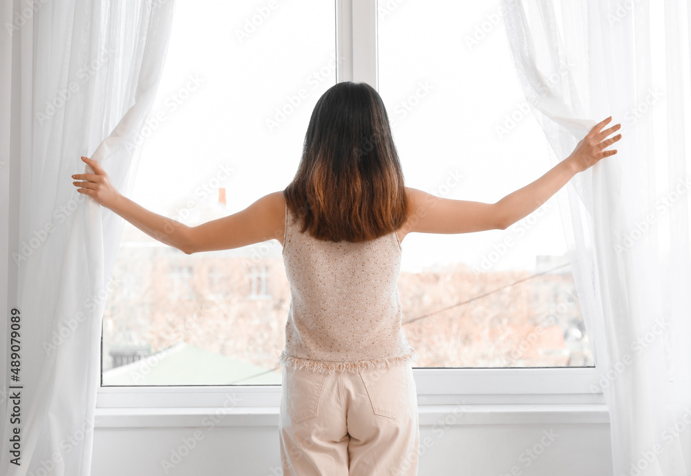 Pretty young Asian woman opening curtains in room Pretty young Asian woman opening curtains in room