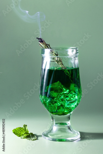 a green drink with ice and a smoldering sprig of rosemary in a glass, mint on a green background. Tarragon, absinthe, green cocktail with smoke
