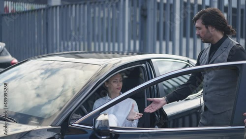Elegant man opening car door for beautiful young woman in urban city outdoors. Portrait of gallant Caucasian businessman helping successful businesswoman walking out the automobile in slow motion