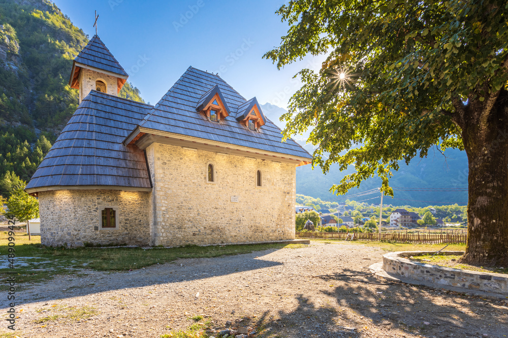 A stone church with a wooden roof in the village of Theth, Albania. Theth is a national park. There is a wooden rustic cross on the roof of the church. The village of Theth is surrounded by mountains.