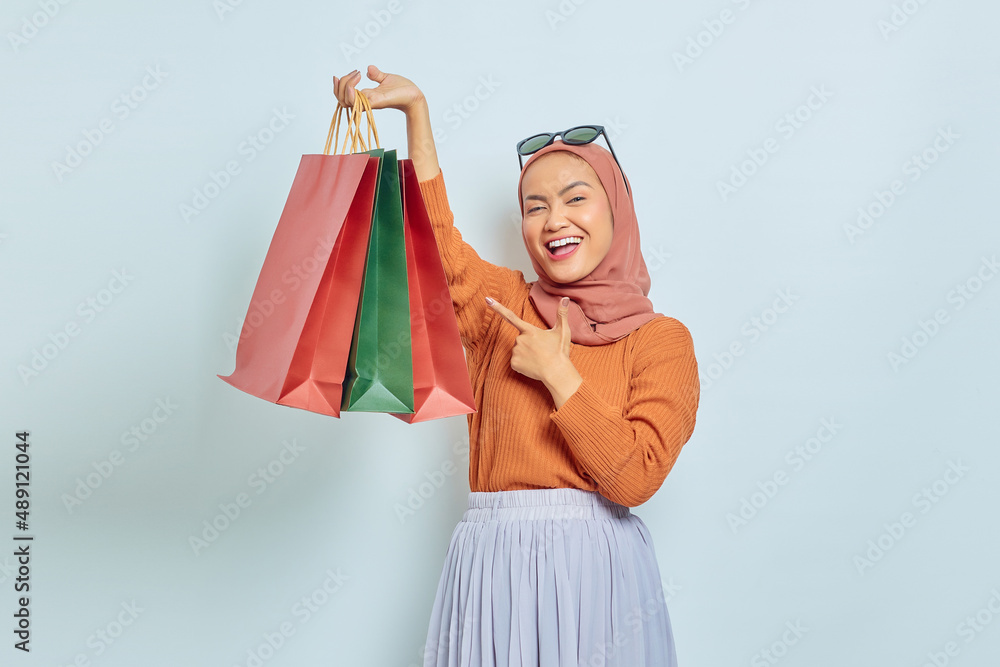 Cheerful beautiful Asian Muslim woman in brown sweater and glasses pointing to shopping bag with finger isolated over white background. World Consumer Rights Day Concept Cheerful beautiful Asian Muslim woman in brown sweater and glasses pointing to shopping bag with finger isolated over white background. World Consumer Rights Day Concept