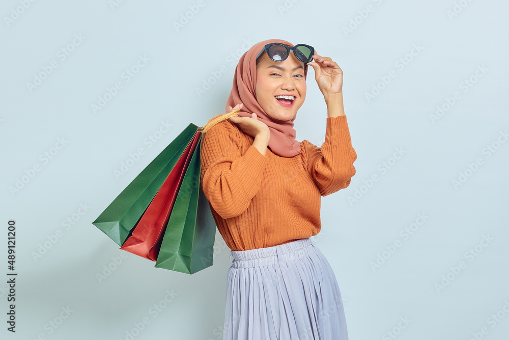 Portrait of excited young Asian Muslim woman in brown sweater sunglasses holding package bag with purchases after shopping isolated on white background. World Consumer Rights Day Concept