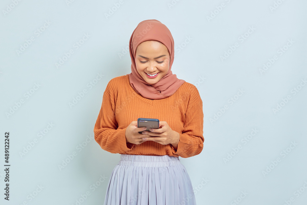 Portrait of smiling young Asian Muslim woman in brown sweater hijab, using mobile phone to send message isolated on white background. Muslim lifestyle concept Portrait of smiling young Asian Muslim woman in brown sweater hijab, using mobile phone to send message isolated on white background. Muslim lifestyle concept