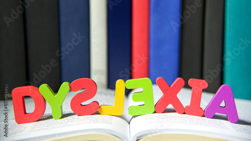 Bright colorful wooden toy blocks on an open book spelling dyslexia. Learning challenges disabilities. S, L and E are backwards. Hard bound books lined up in background photo