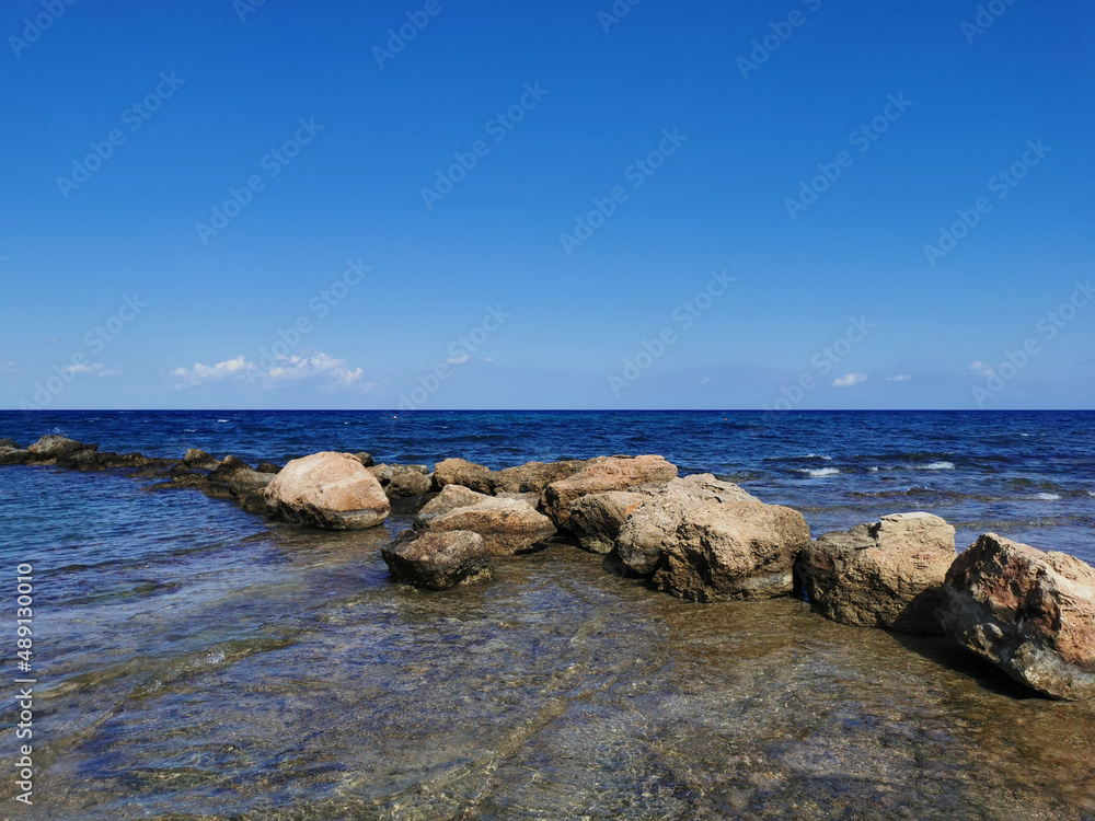 The coast of the Mediterranean Sea, waves, clear water, a stone ridge against a blue sky with clouds.