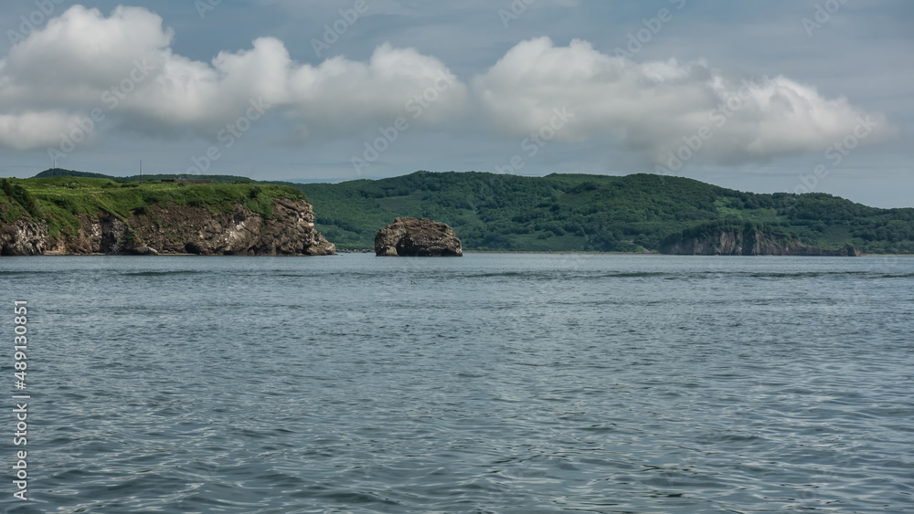 The picturesque coast of Kamchatka. Rocks and green hills against a blue sky with clouds. Ripples on the surface of the Pacific Ocean. Avacha Bay The picturesque coast of Kamchatka. Rocks and green hills against a blue sky with clouds. Ripples on the surface of the Pacific Ocean. Avacha Bay
